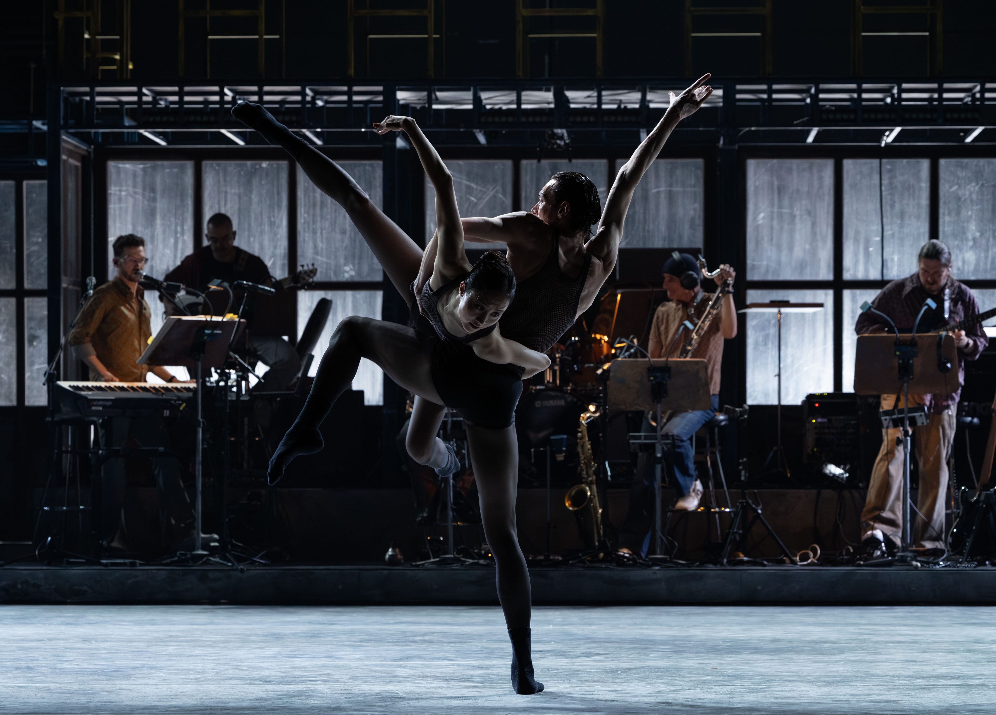 Zwei Tänzer*innen vor einem Bühnenbild, das eine Industriekulisse darstellt.